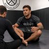 Tattooed BJJ athlete in Reedot Sports spider web rash guard and black shorts seated in grappling position on mats